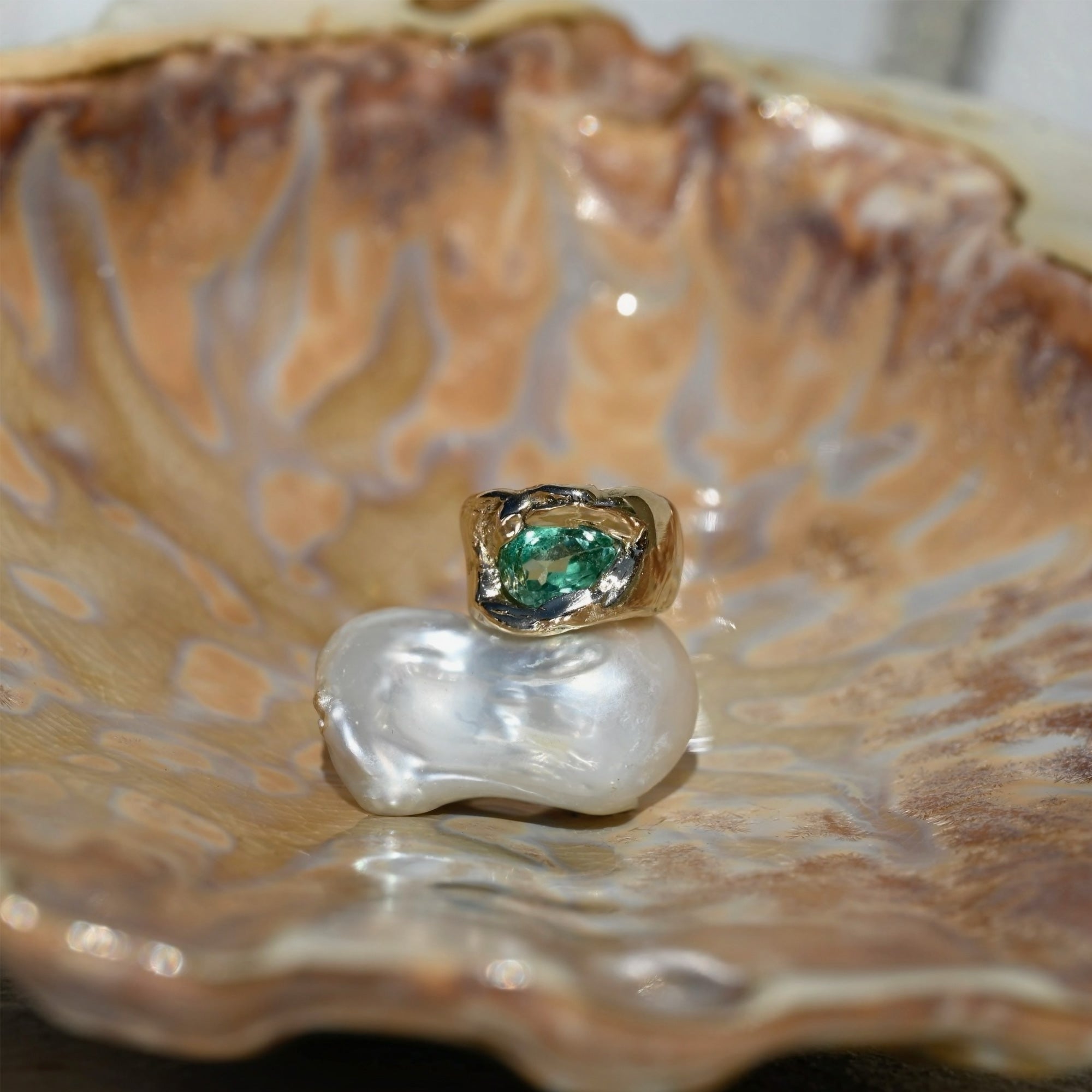 Gold ring with a green gemstone on a white pearl, set against a large shell background.